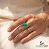 Hand wearing a gold ring with an emerald cross pendant on a beige fabric background, with 'SM Gems India' branding.