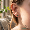 Close-up of an ear wearing green gemstone earrings with a blurred outdoor background.