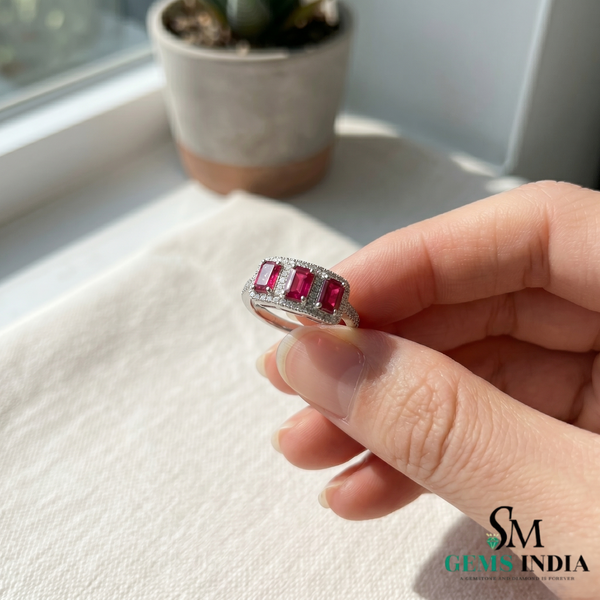 Hand holding a ring with red gemstones on a white surface with a plant in the background